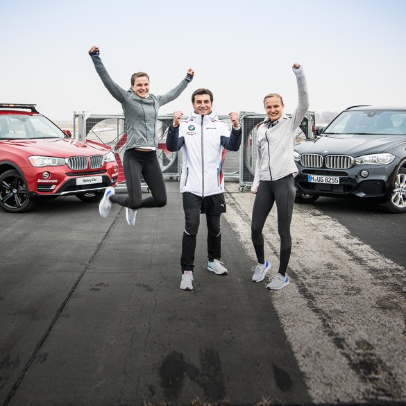 Bruno und Hahnertwins jubelnd vor zwei BMW Fahrzeugen auf Straße.